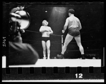 A wrestler strides toward Gorgeous George who stands near a corner of the ring with his hands on his chest where he had received a blow in the previous maneuver in the wrestling match